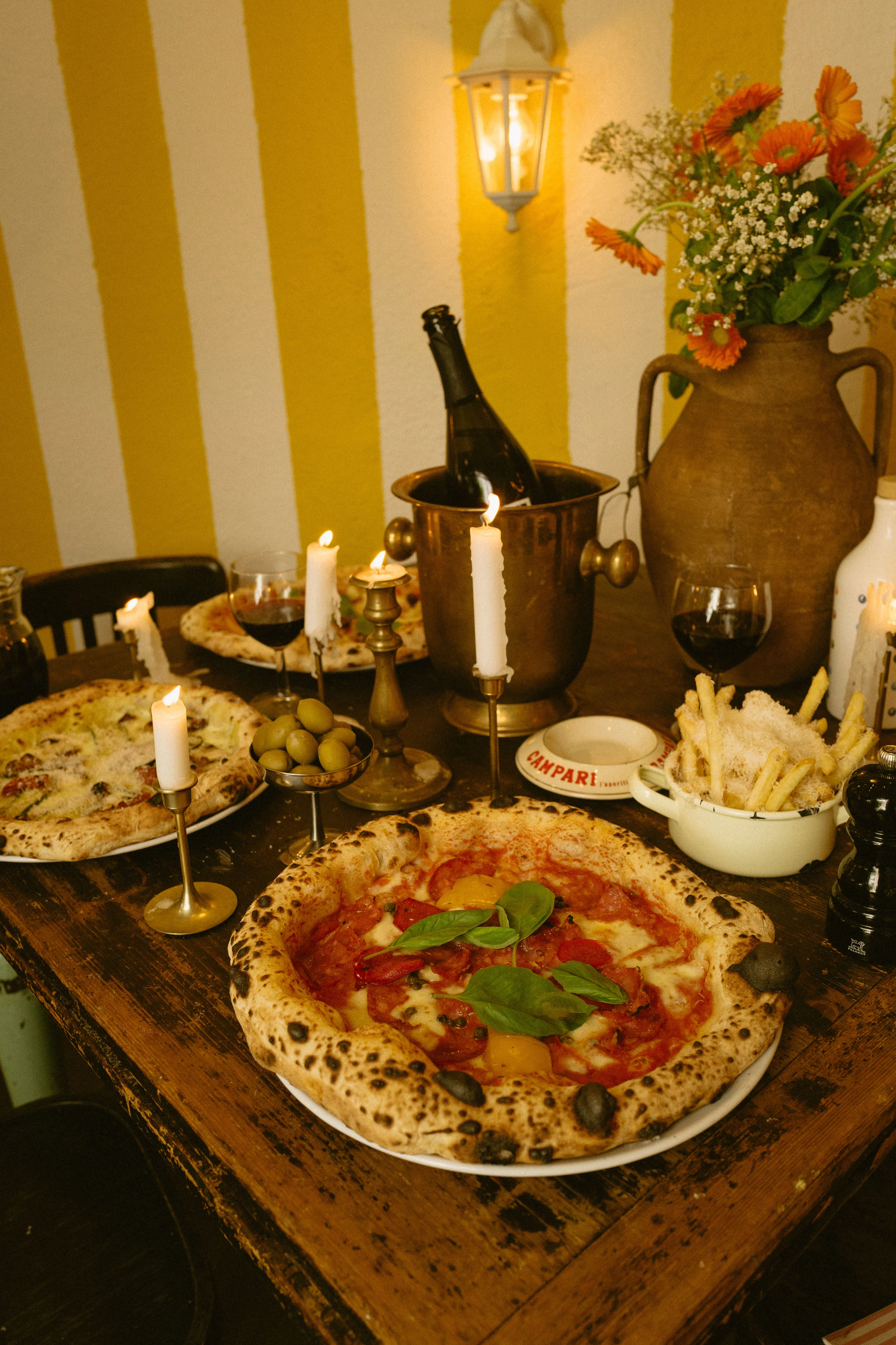 Ein gedeckter Holztisch in einem italienischen Restaurant mit Pizza, Pommes frites, Wein und brennenden Kerzen vor einer gelb-weiß gestreiften Wand.